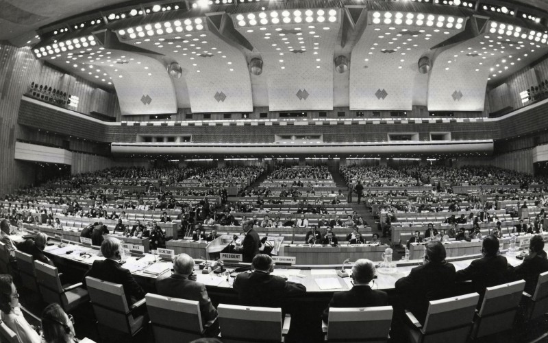 Black and white wide-angle shot of cavernous building with people seated at large table facing banks of stadium seats.