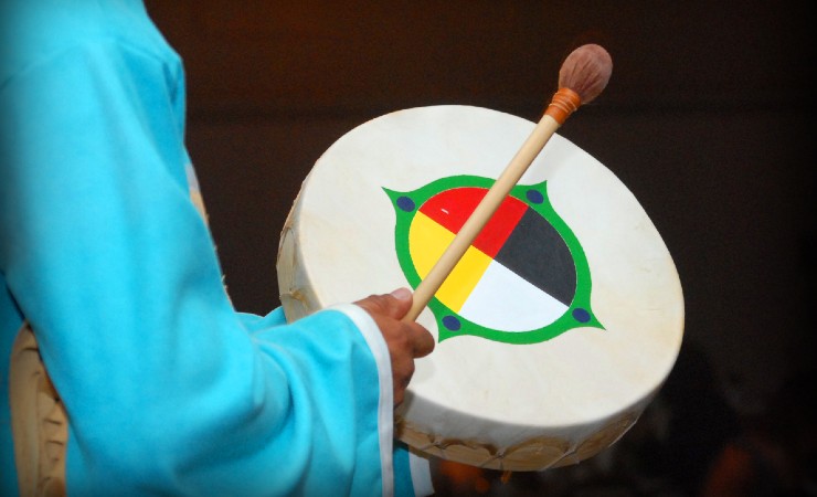 Person in blue robe uses drumstick to beat hand-held drum with art on skin