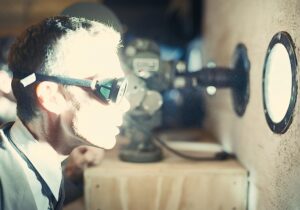 Actor wearing goggles views enormously bright light shining in through porthole-type window