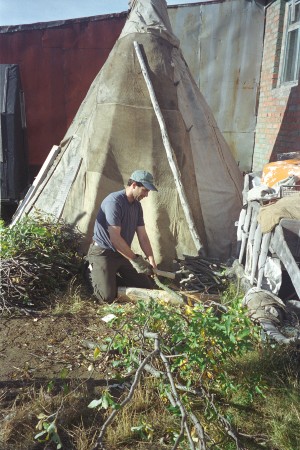 Man in T-shirt cutting branches in front of conical tent