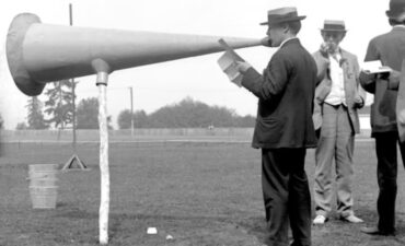 Black and white photo of man from circa 1900 speaking into giant megaphone on a stick