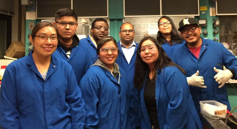 Group shot of eight students in blue lab coats and safety glasses