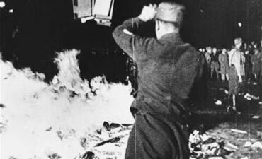 Black and white photo of man in uniform throwing books onto large roaring bonfire