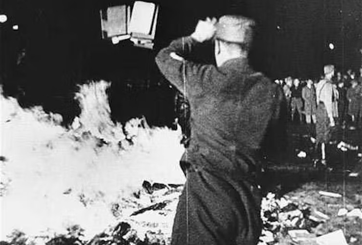 Black and white photo of man in uniform throwing books onto large roaring bonfire