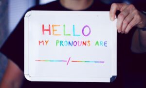 Close-up photo of employee holding whiteboard with "Hello, my pronouns are ____/_____" written in rainbow color markers.