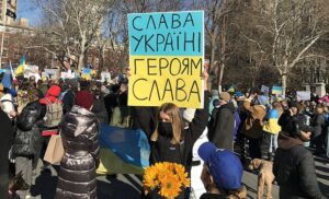 Blonde woman wearing mask holds a sign above her head depicting the Ukrainian flag and reads "glory for Ukraine". She is at a gathering of people protesting the Russian invasion, and is stands in a crowd of people with Ukrainian flags and signs.
