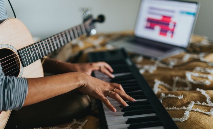 Close-up photograph of a musician playing a keyboard with a guitar in their lap and an open laptop sitting in the background.