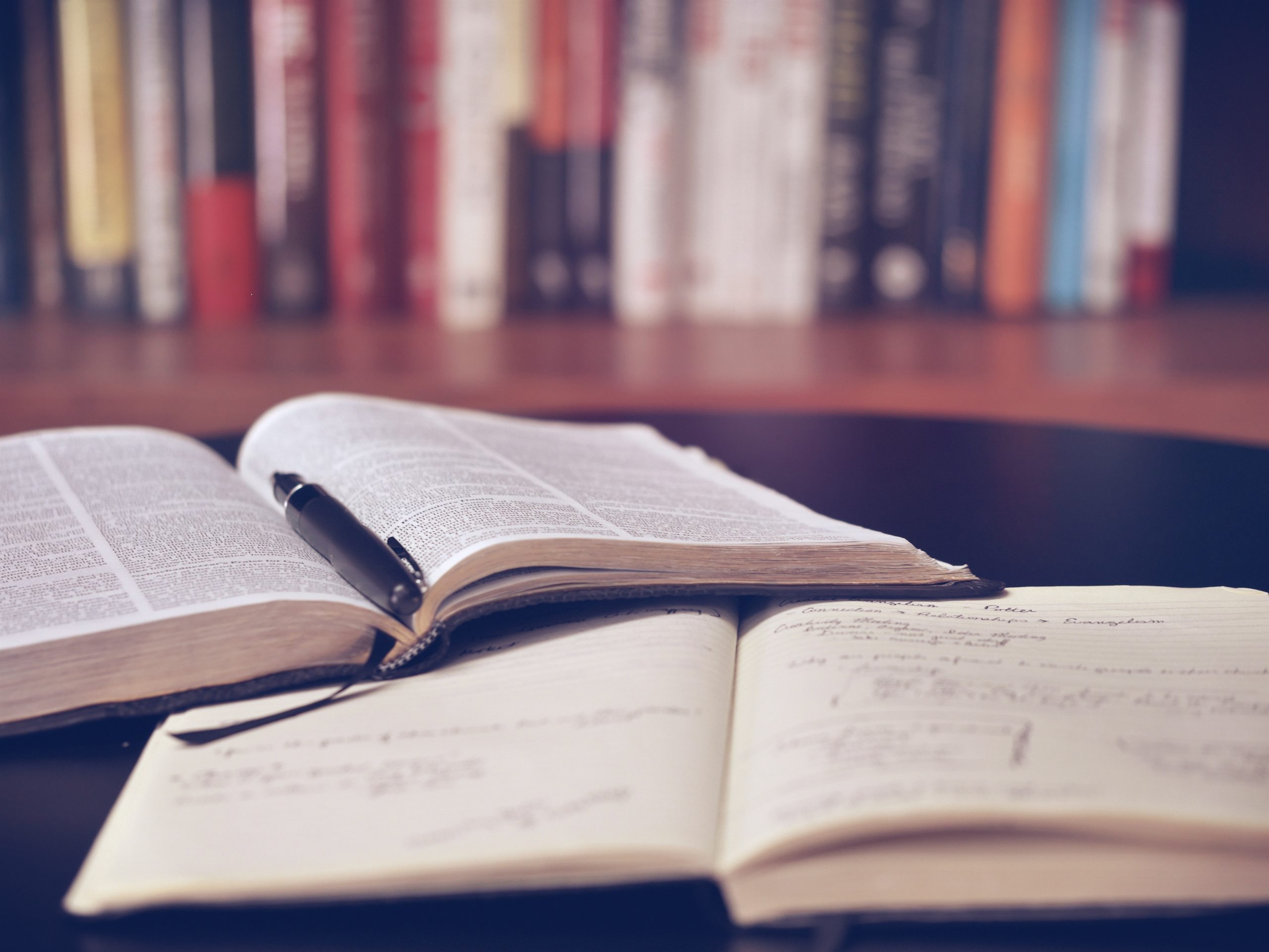 Two books lie open on a table with a pen on top of one, with a line of book spines visible in the background.