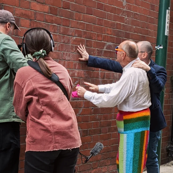 Man with camera and woman with headset on film two men facing a brick wall.