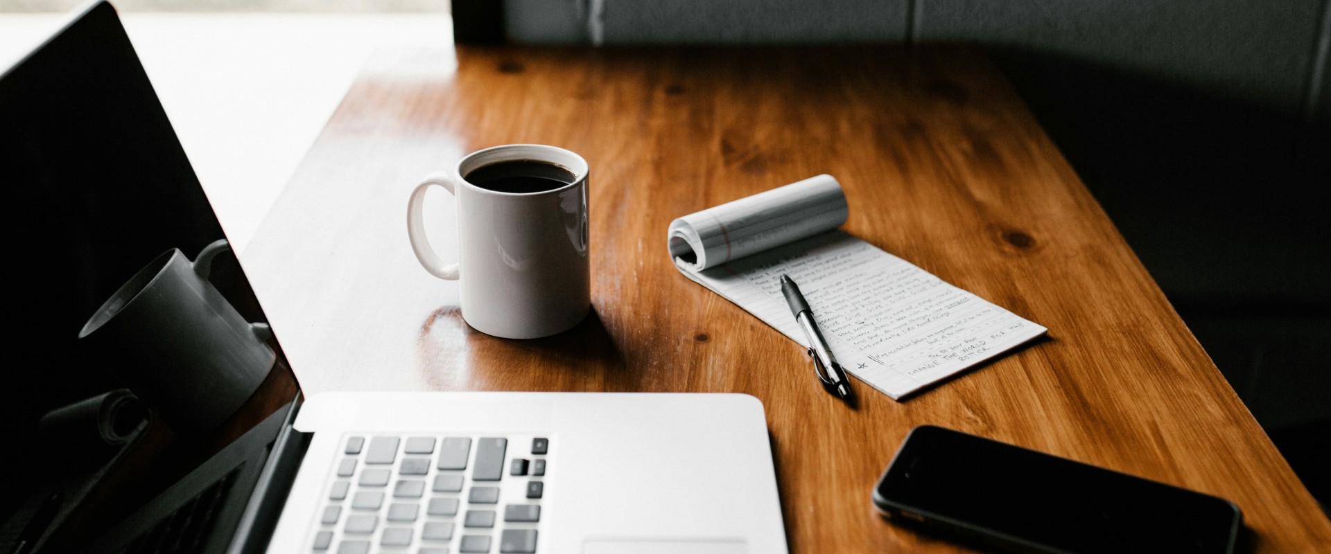 Wooden desk with coffee cup, pad of paper, laptop and phone