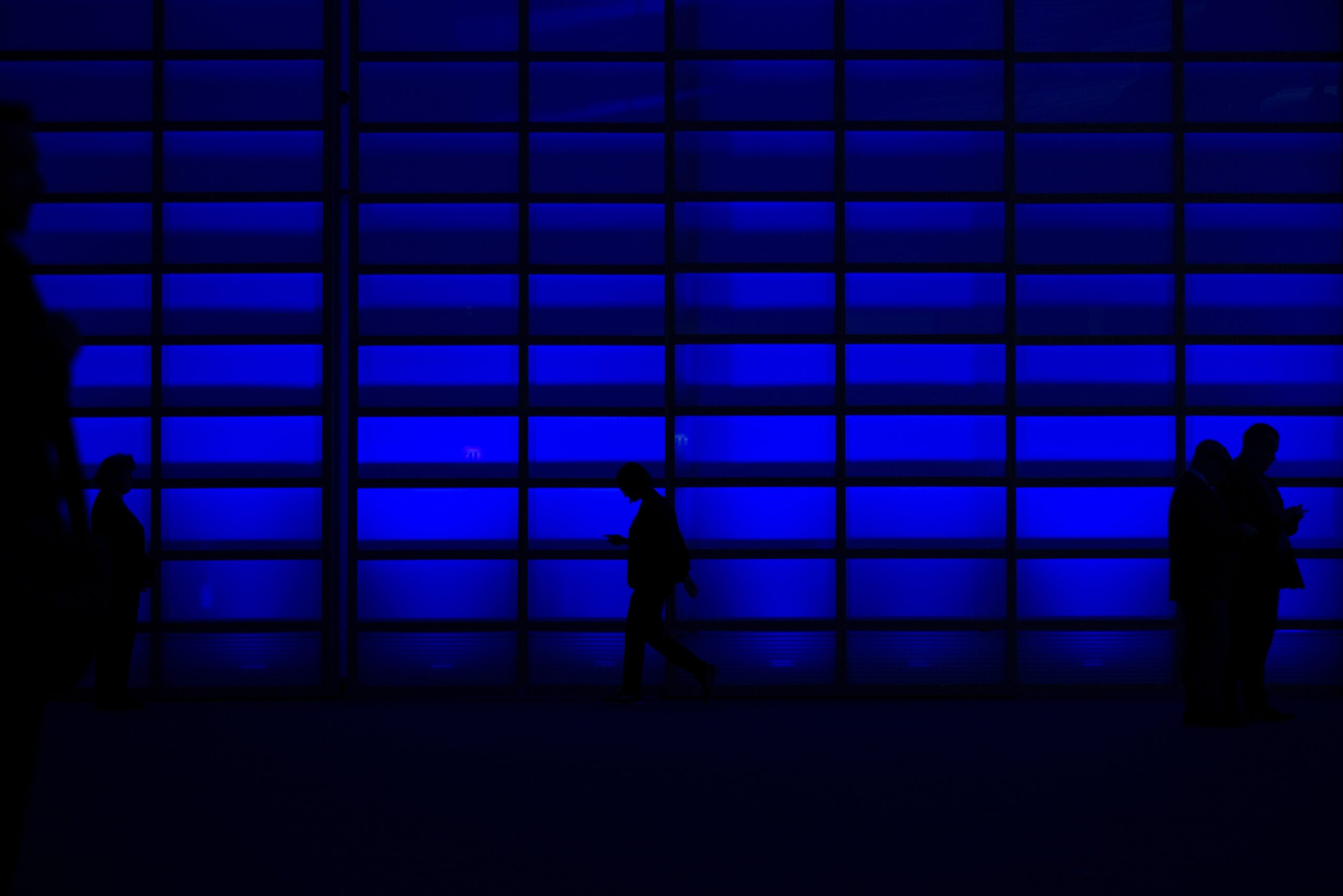 Silhouetted figures pass in front of a dark blue background looking down at mobile phones.